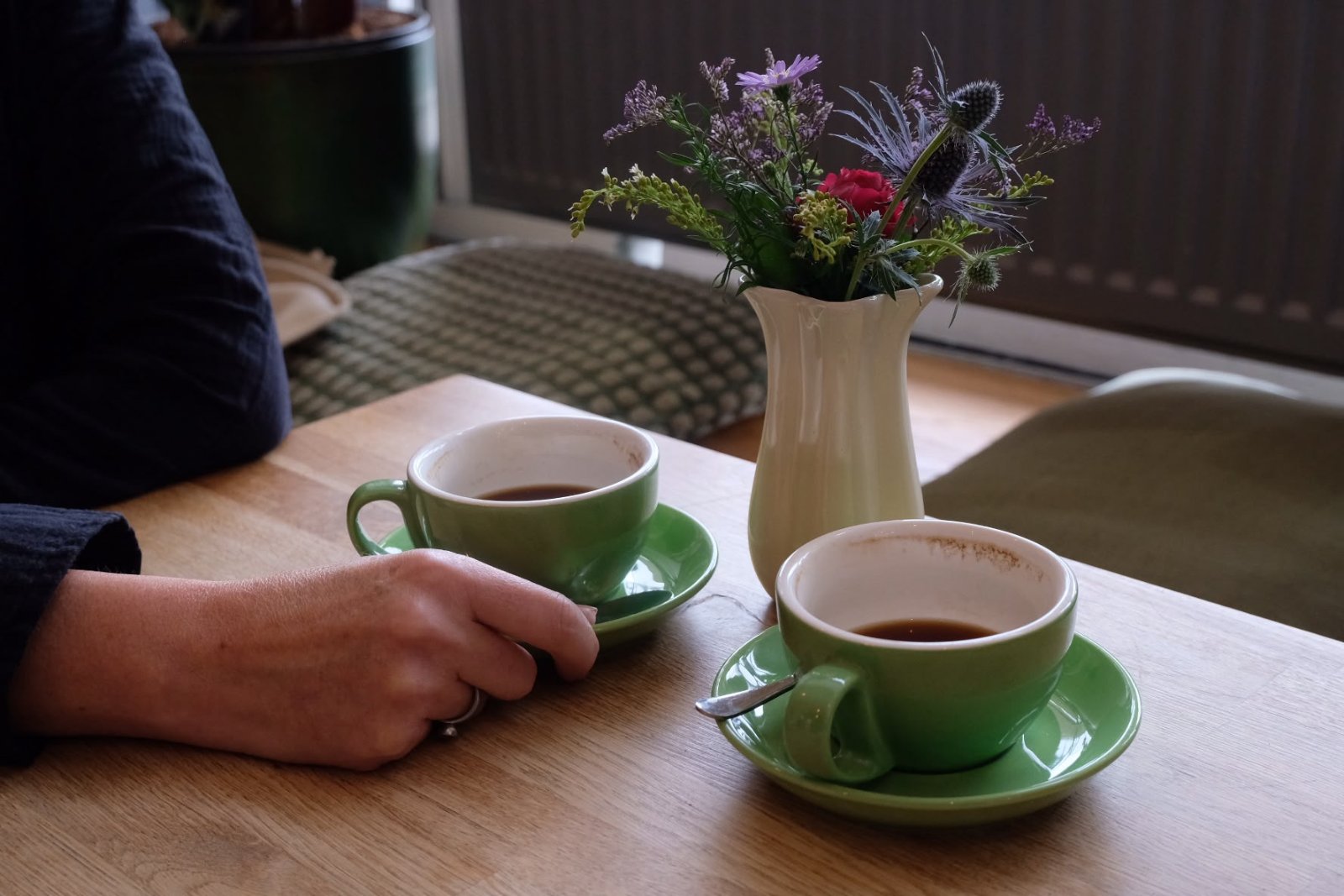 Twee groene kopjes koffie en een vaasje veldbloemen op een houten tafel; een hand rust naast het ene kopje